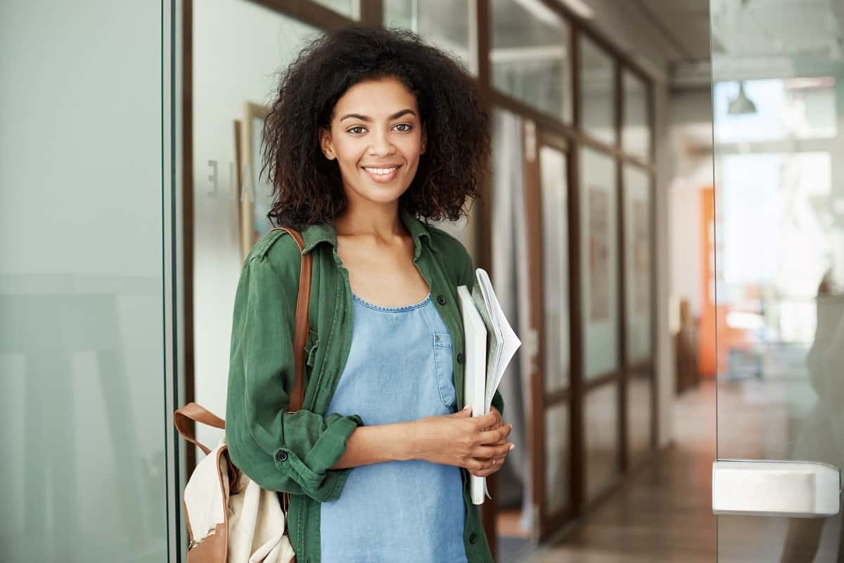 A girl standing while holding some books in her hand and a bag on her shoulder