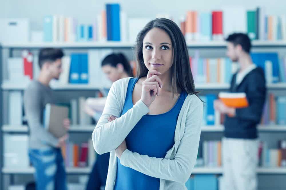 a female student standing and thinking in a library with 3 students in the background with books